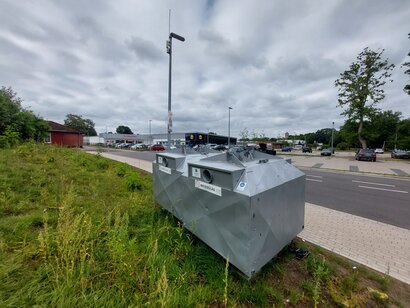 Ein Glascontainer steht auf der Wiese neben der Helga-Mohrhoff-Straße
